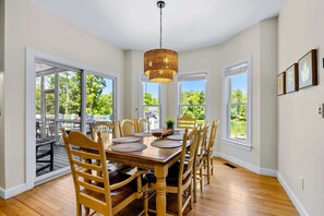 A dining room with a wooden table, chairs, a hanging light fixture, and large windows.