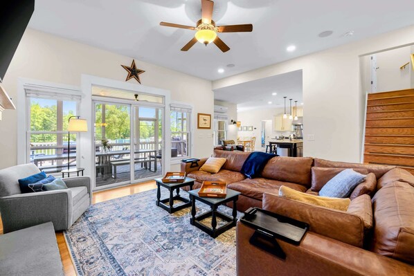 A spacious living room with a brown leather sofa, a gray armchair, and a patterned rug. There is a ceiling fan, a star decoration, and a view of the outdoors through large windows.