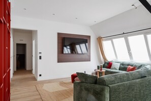 Un salon moderne avec un grand téléviseur à écran plat fixé au mur, un canapé vert agrémenté de coussins rouges, une petite table d’appoint et un plancher en bois.