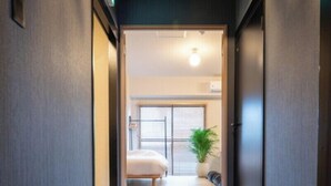 Ein Flur, der zu einem Zimmer mit einem Bett, einer Pflanze und einem Fenster mit Jalousien führt.