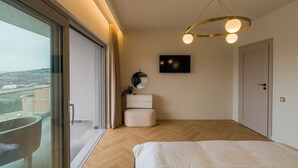 A bedroom with a large window, a bed, a bedside table, a mirror, and a wall-mounted TV.