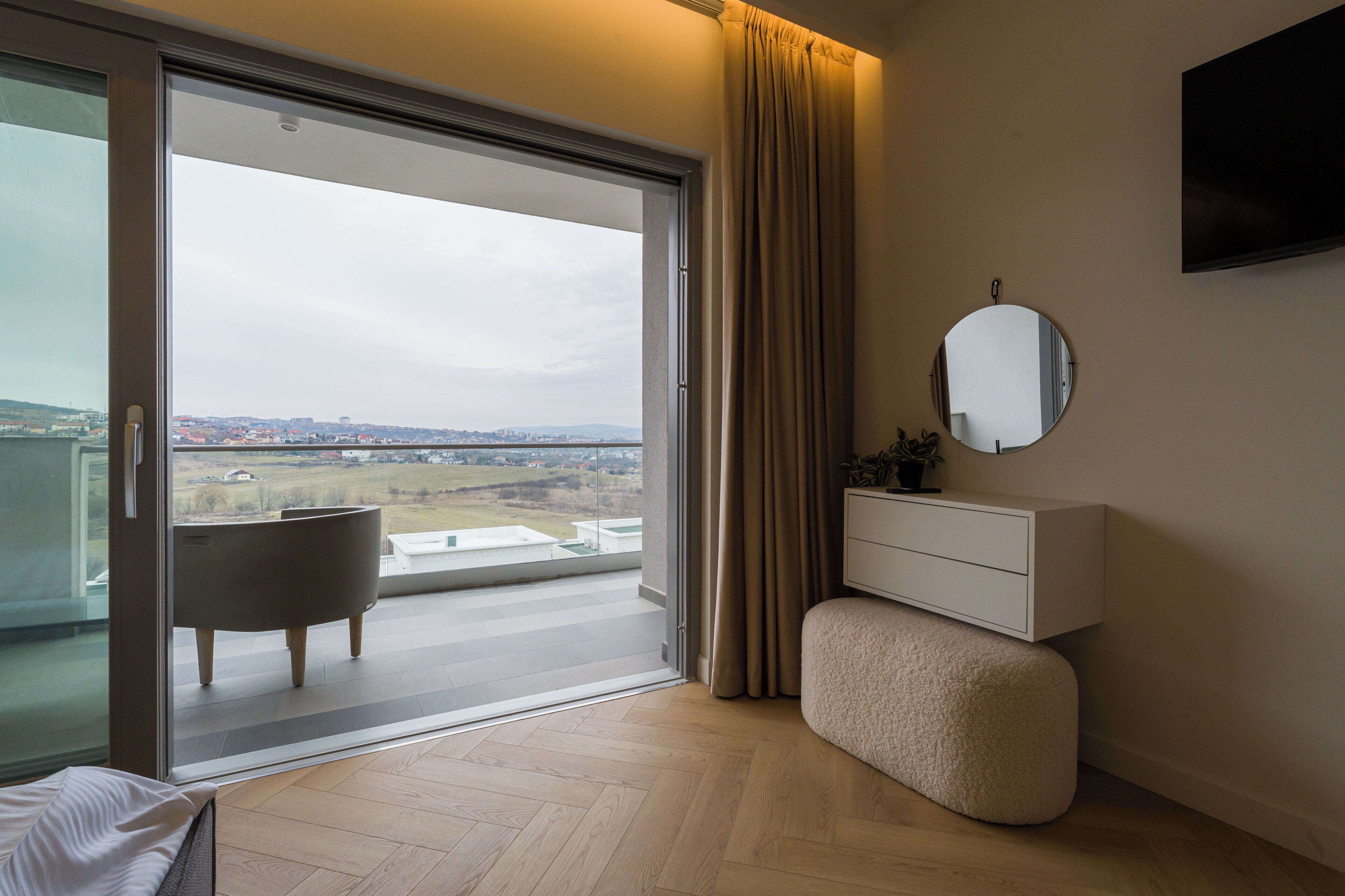 Une chambre à coucher moderne avec une porte-fenêtre coulissante donnant sur un balcon, un miroir rond, une commode blanche et un pouf capitonné.