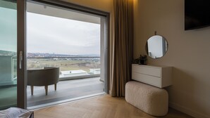 A modern bedroom with a sliding glass door leading to a balcony, a round mirror, a white dresser, and a tufted ottoman.