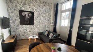 Un salon moderne avec une table de salle à manger ronde en bois, un canapé noir, un tableau représentant un éléphant accroché au mur, et un coin cuisine doté d’appareils électroménagers encastrés.