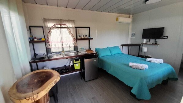 A compact room with a bed, a desk with a sink, a small refrigerator, and a television on the wall.