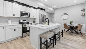 A modern kitchen with white cabinetry, stainless steel appliances, a central island with bar stools, and a dining area with a round table and chairs. - Freshly renovated 2 story house near downtown Indy (Indianapolis)