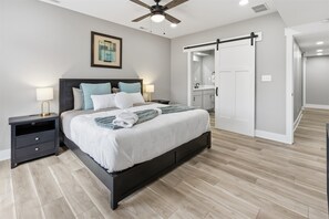 Ein Schlafzimmer mit Bett, Nachttischen, Deckenventilator, einem gerahmten Bild, einer Schiebetür und einem durch die Tür sichtbaren Badezimmer.