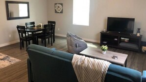 A living room with a blue sofa, a coffee table with a flower vase, a TV stand, and a dining area with chairs and a table.