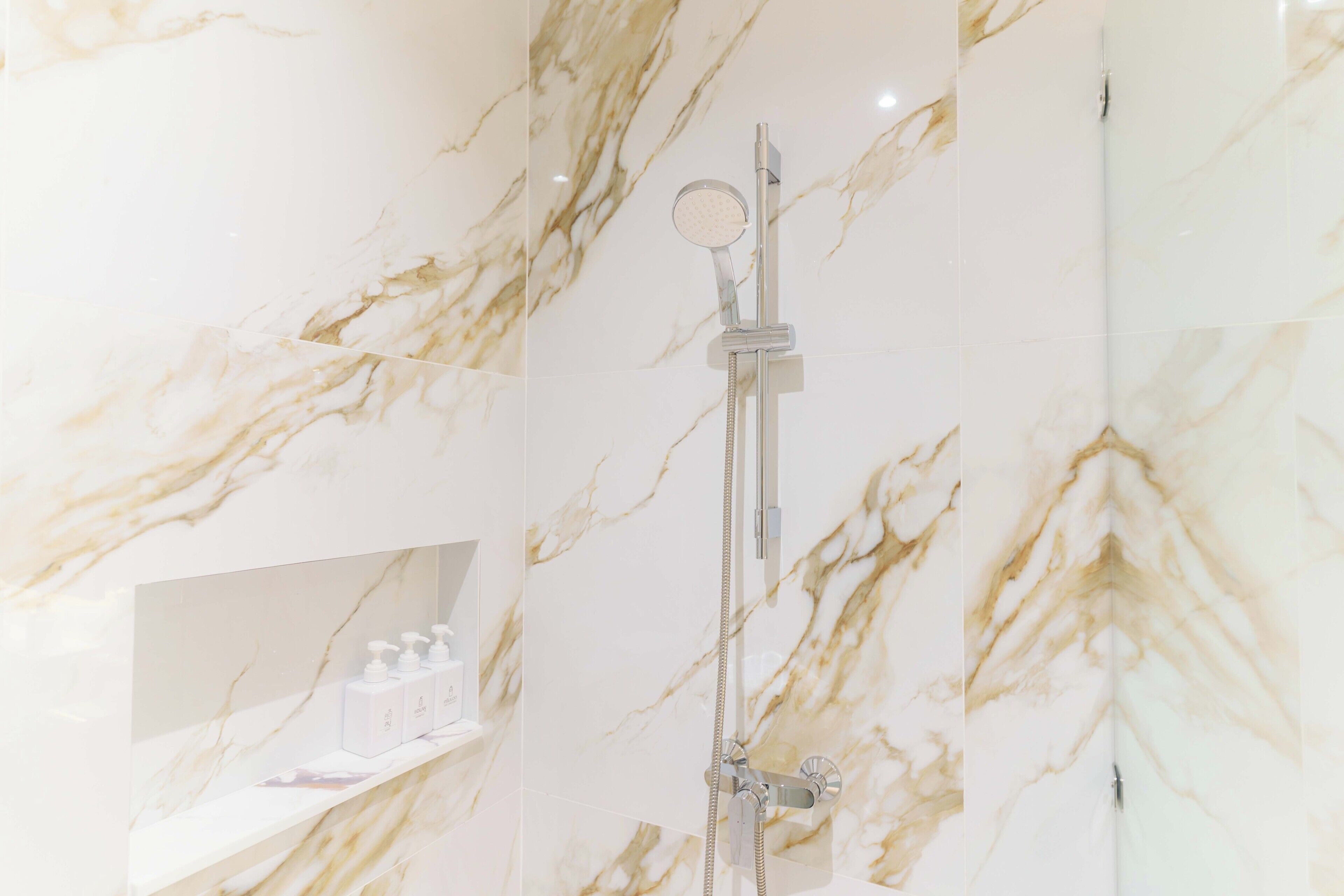 A bathroom with a marble wall, a handheld showerhead, and a shelf with toiletries.