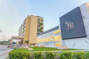 A hotel entrance with a large sign for 'Sakhonburi Cozy Hotel,' a modern building with a flat roof, and a clear sky.
