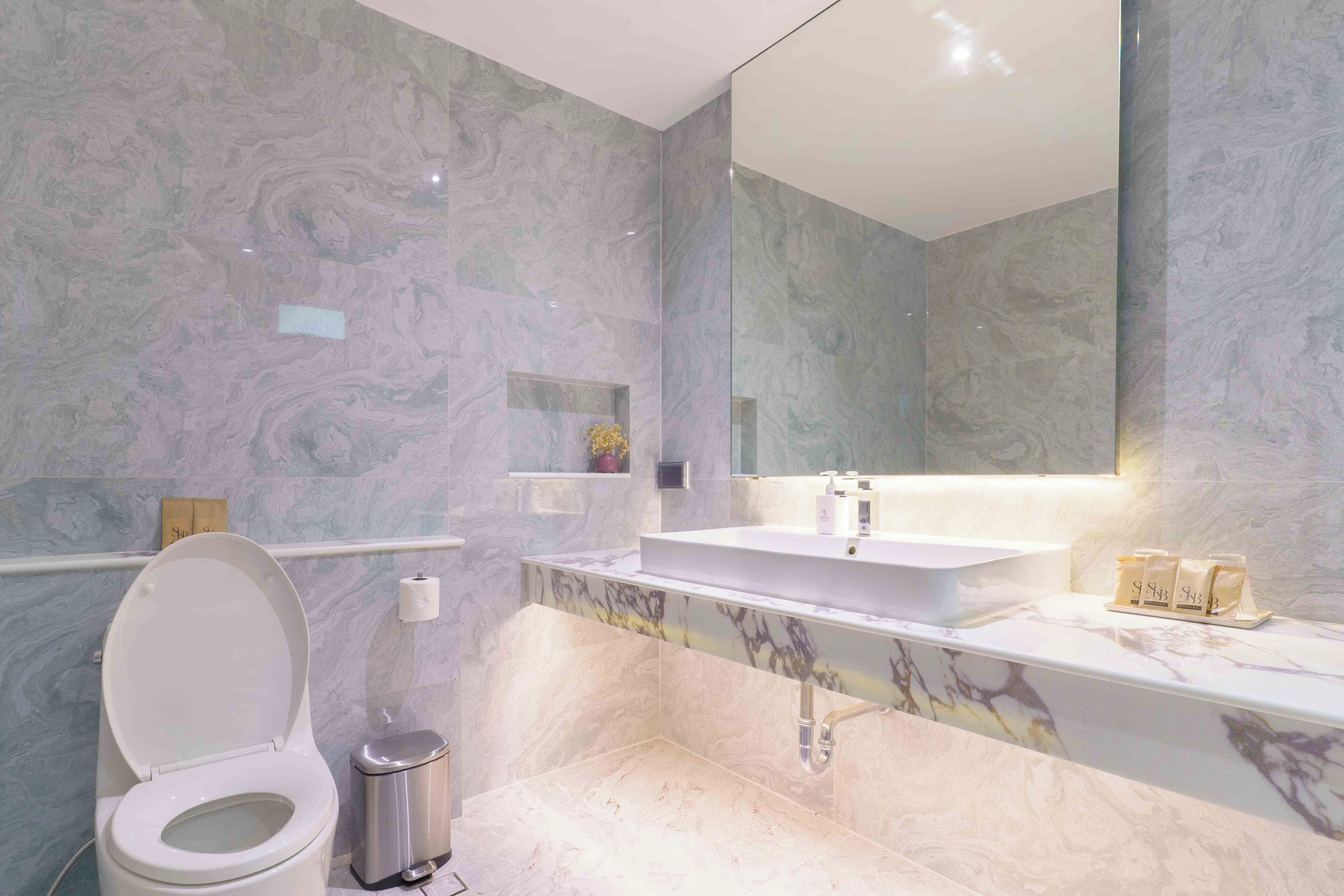 Un baño con inodoro, lavamanos, espejo, paredes de mármol y un cubo de basura.
