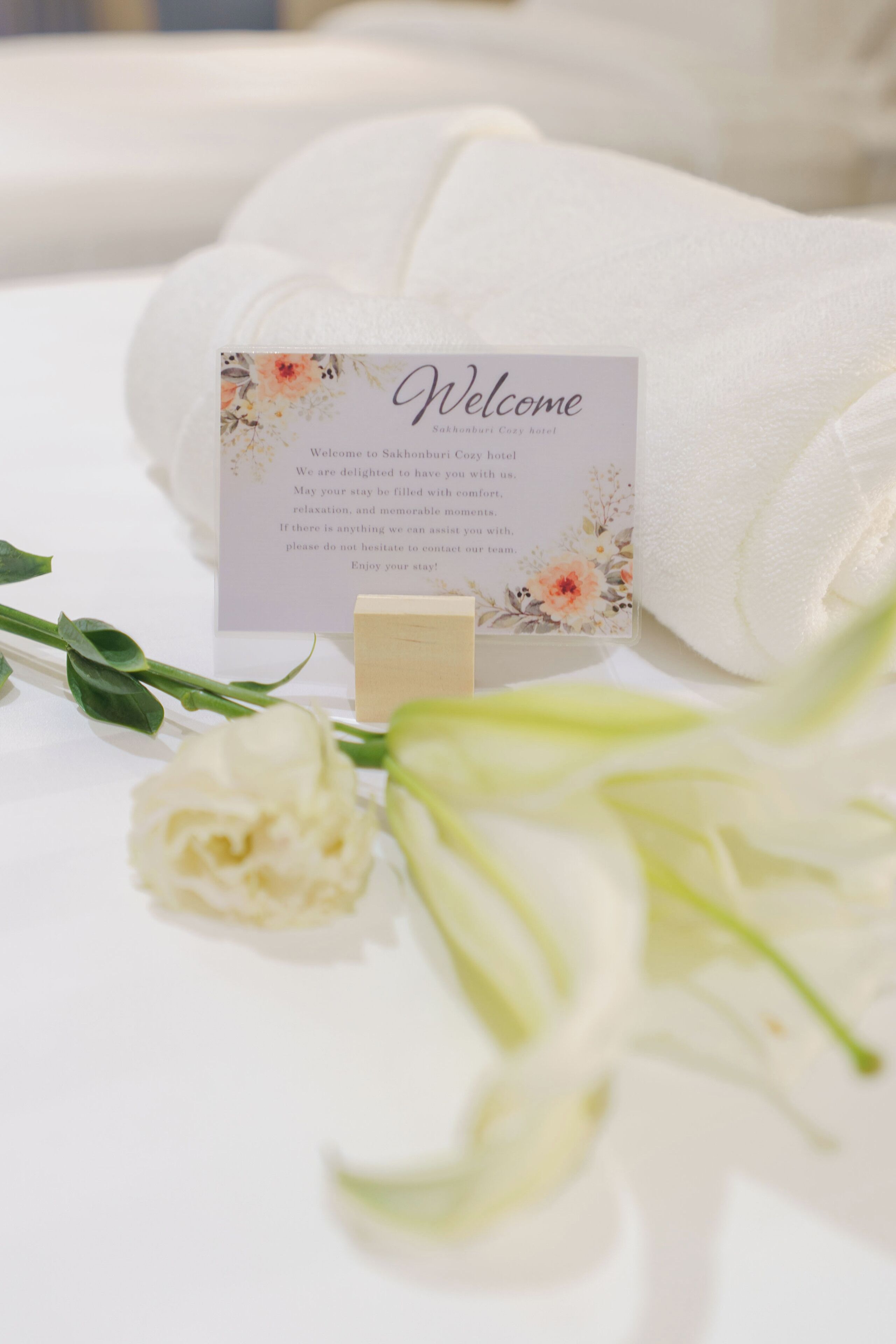 A welcome card on a bed with a floral design, a white rose, and a green lily.