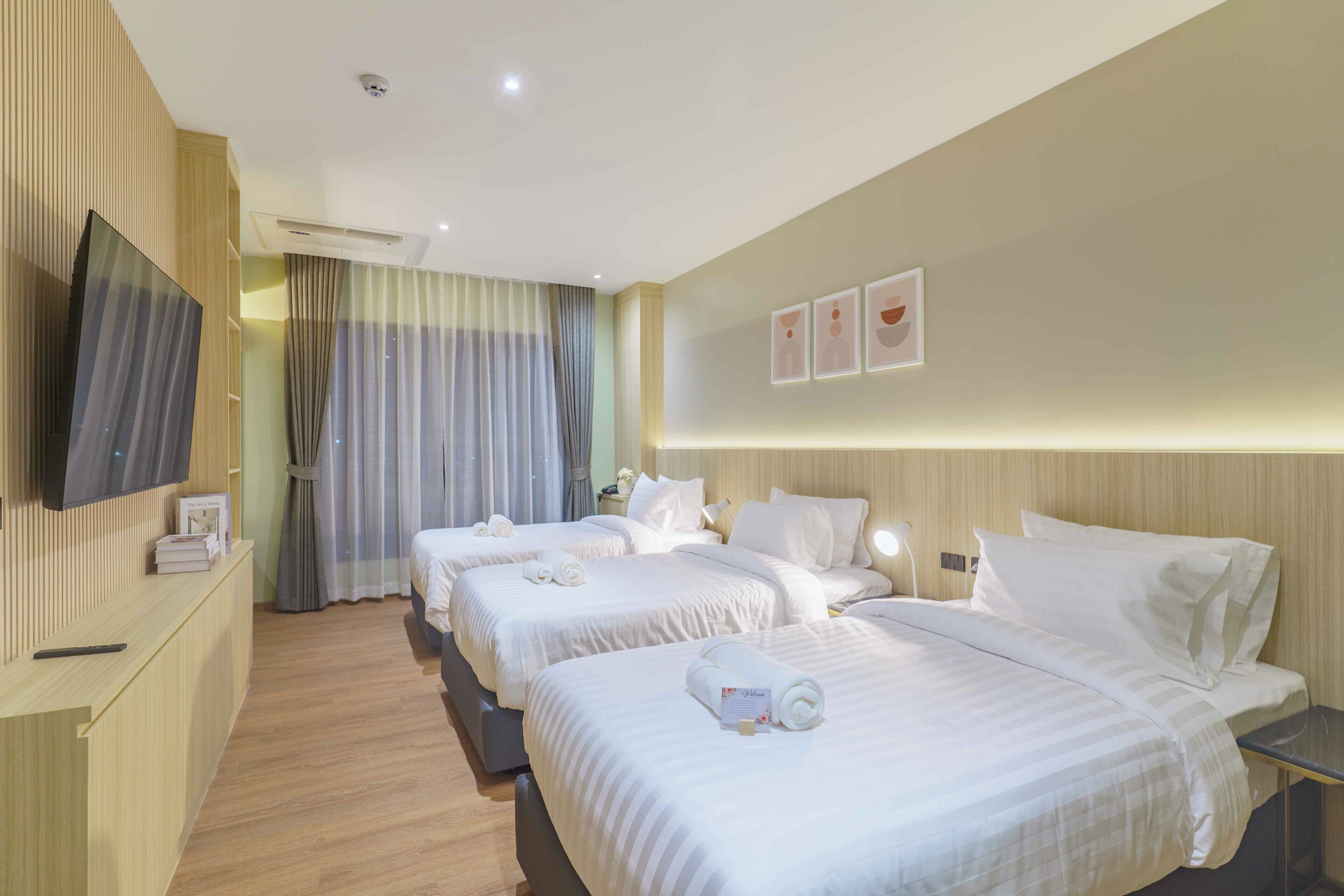 A hotel room with three beds, a TV on the wall, a desk, and a window with curtains.