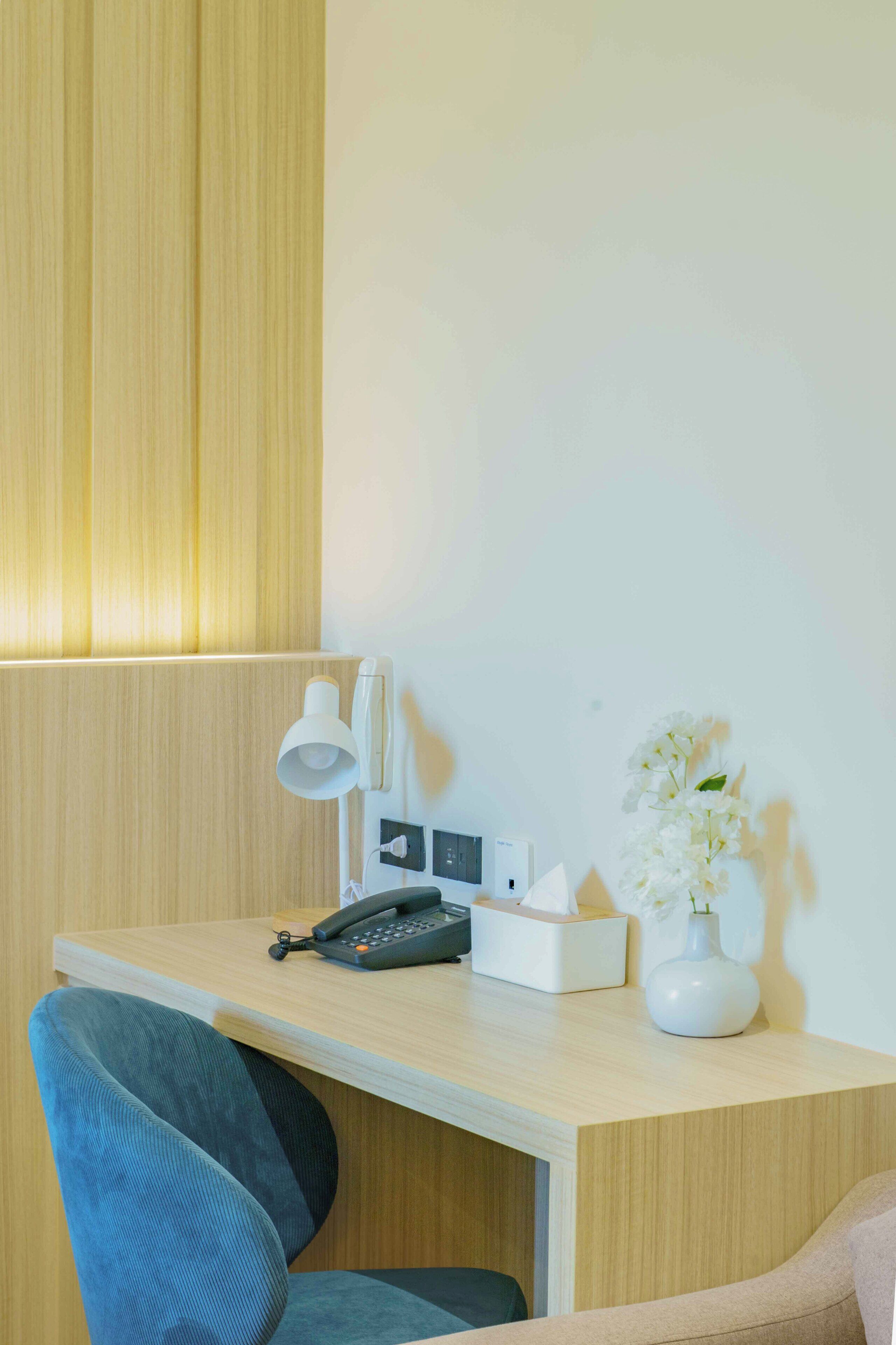 A hotel room with a wooden desk, a telephone, a lamp, a tissue box, and a vase with flowers.