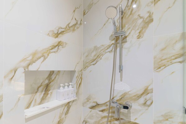 A bathroom with marble walls, a shower, and a shelf with toiletries.
