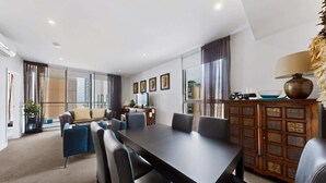A modern hotel room with a large dining table, comfortable seating, a wooden cabinet, and large windows. - Skyline 2-Bed Apartment Near Metro North Ryde (North Ryde)