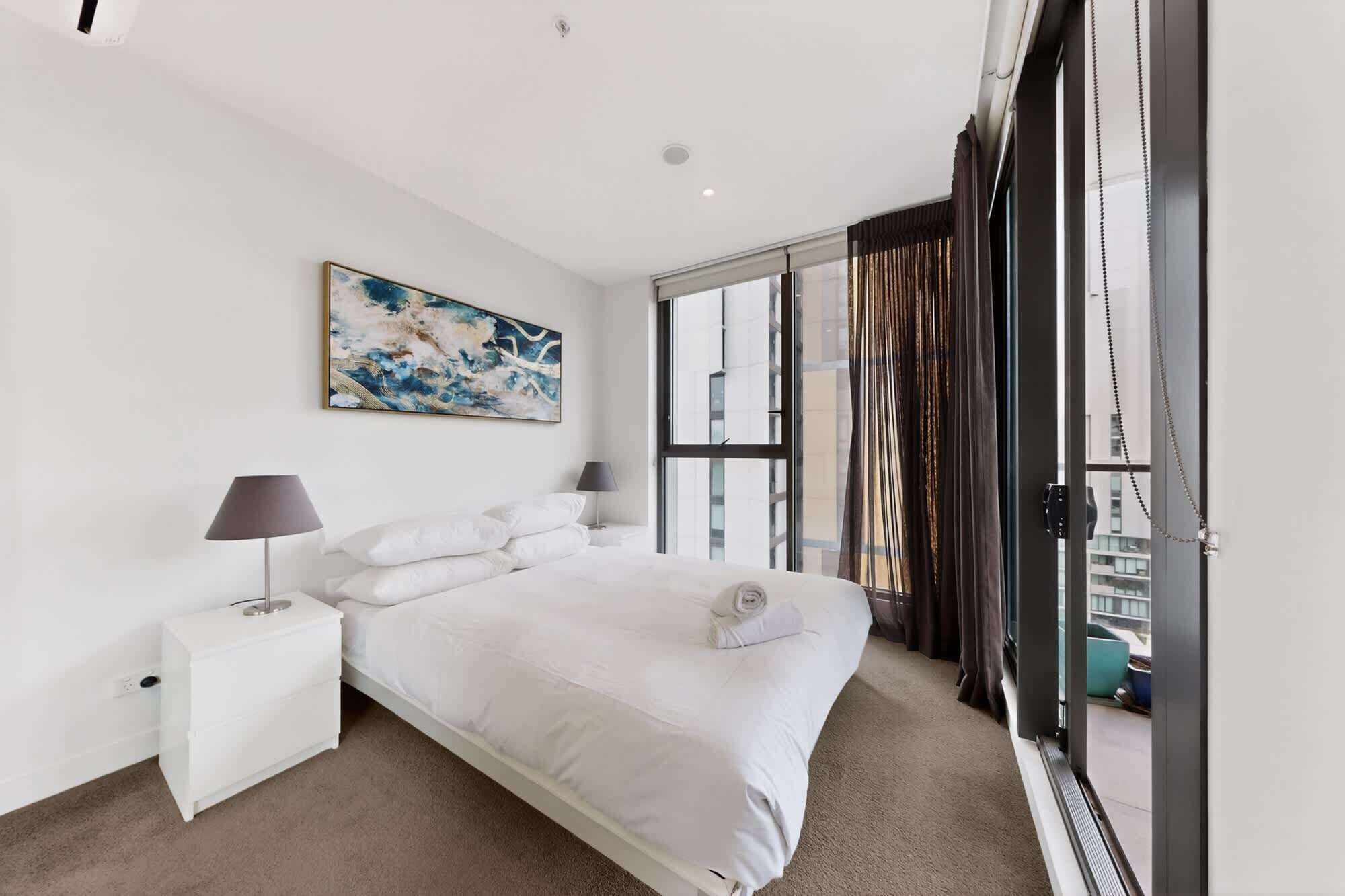 A modern bedroom with a large bed, a bedside table, a nightstand, a window with curtains, and a city view.