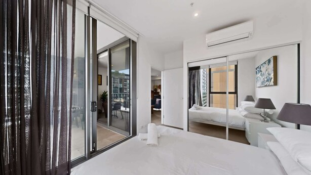 A modern hotel room with a large bed, a sliding glass door to the balcony, a separate seating area, and a large mirror. - Skyline 2-Bed Apartment Near Metro North Ryde (North Ryde)