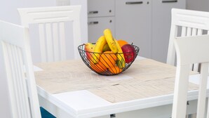 A white dining table with a woven placemat, surrounded by white chairs. A fruit bowl with bananas, oranges, and apples is placed on the table. - Richard Sea View Apartment (Qendër Vlorë)