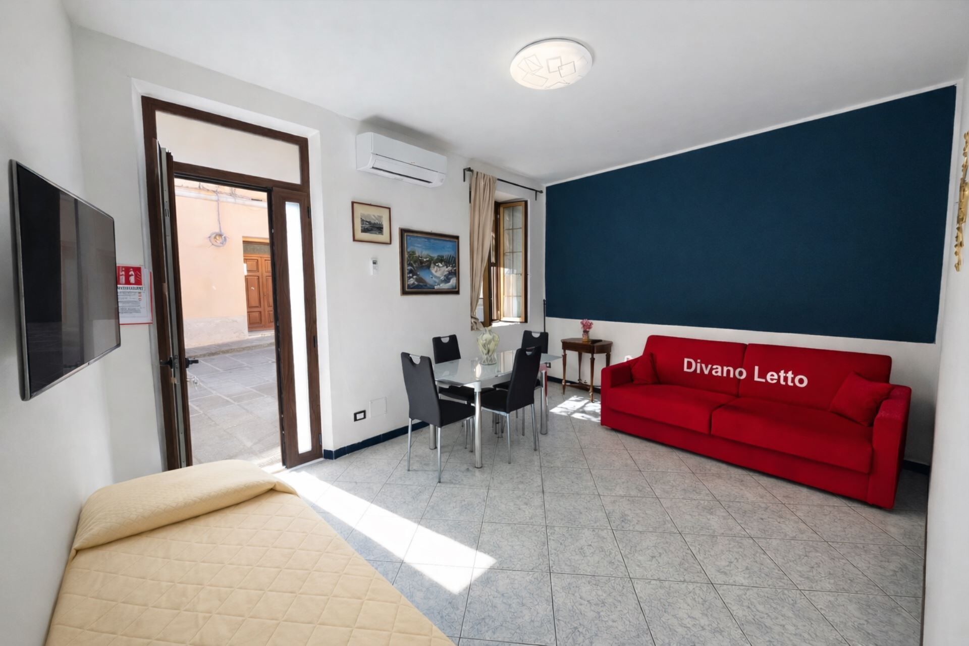 A room with a bed, a red sofa, a dining table with chairs, a TV, and a window with curtains.