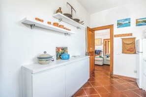 Una cocina con armarios blancos, encimera blanca y estantes con utensilios de cocina. Hay una puerta que conduce a otra habitación.