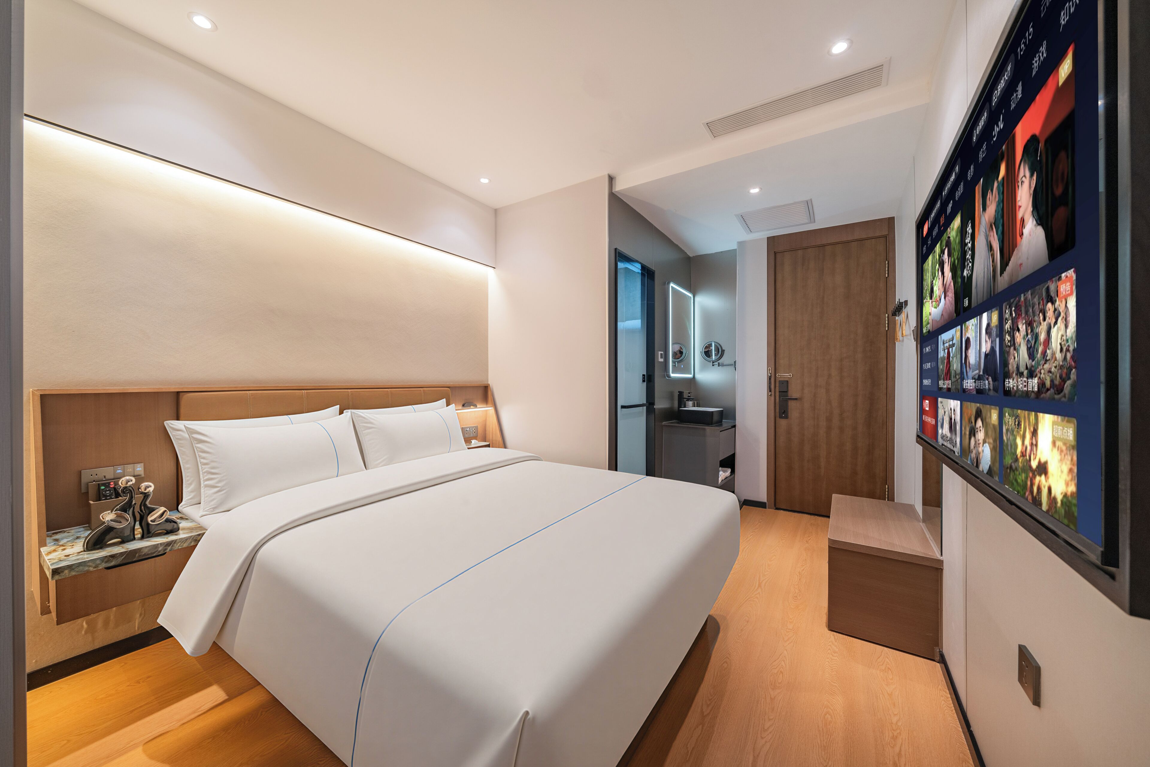 A modern hotel room with a large bed, a bedside table with a phone and charger, a small desk, a built-in TV, and a wooden floor.