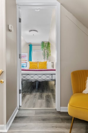 Un pasillo que conduce a un dormitorio con una cama, un sillón amarillo y una lámpara en el techo.