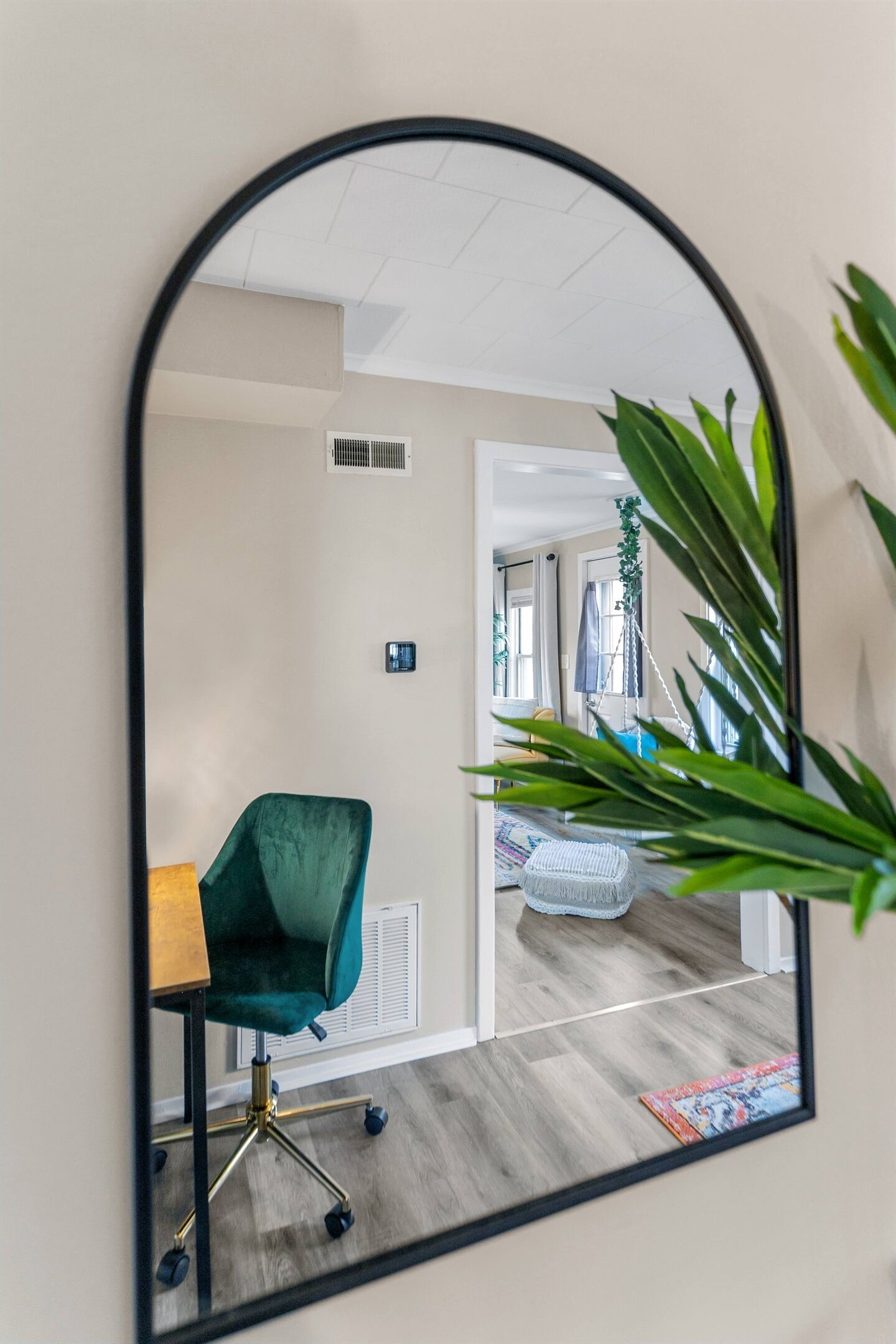 Un miroir reflétant une pièce dans laquelle on peut voir une chaise verte, une petite table, une plante suspendue et une porte donnant sur une autre pièce.