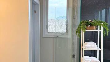 A hallway with a glass door leading to a room, a white shelf with towels, and a potted plant.
