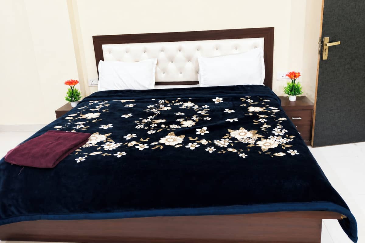 Una cama con colcha floral, dos almohadas blancas, un cabecero de madera oscura, una mesita de noche de madera oscura con una planta, y una puerta con pomo.