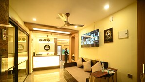 A hotel lobby with a reception desk, a sofa, a ceiling fan, and a wall-mounted TV. - ELORA'S INN (Siliguri)