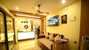A hotel lobby with a reception desk, a seating area with a sofa, a ceiling fan, and a wall-mounted TV. - ELORA'S INN (Siliguri)