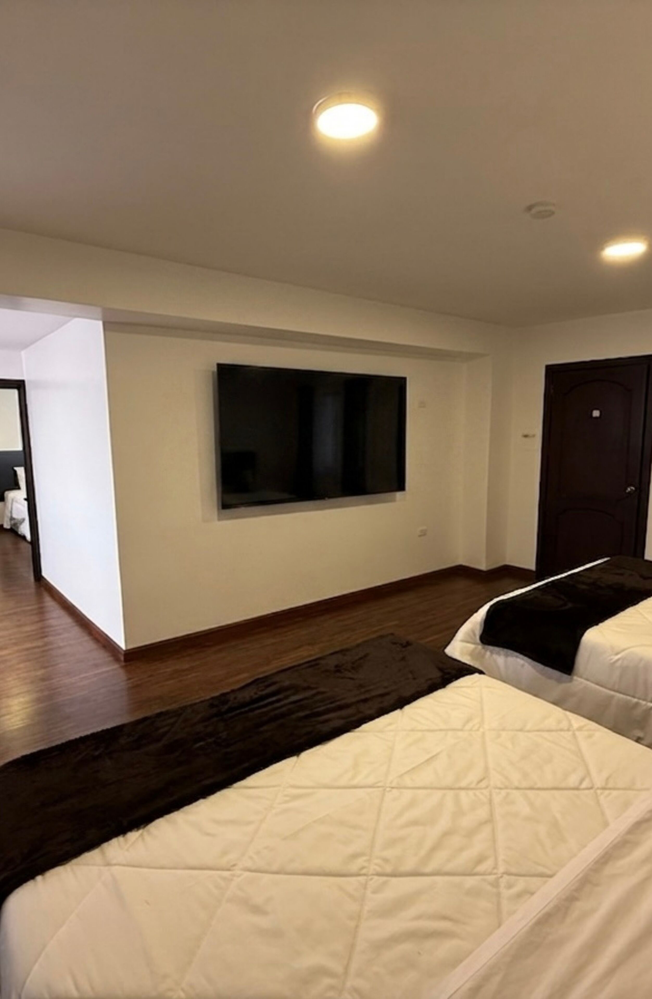 Une chambre d’hôtel dotée d’un grand téléviseur à écran plat fixé au mur, d’un parquet en bois foncé et de deux lits avec une literie blanche.