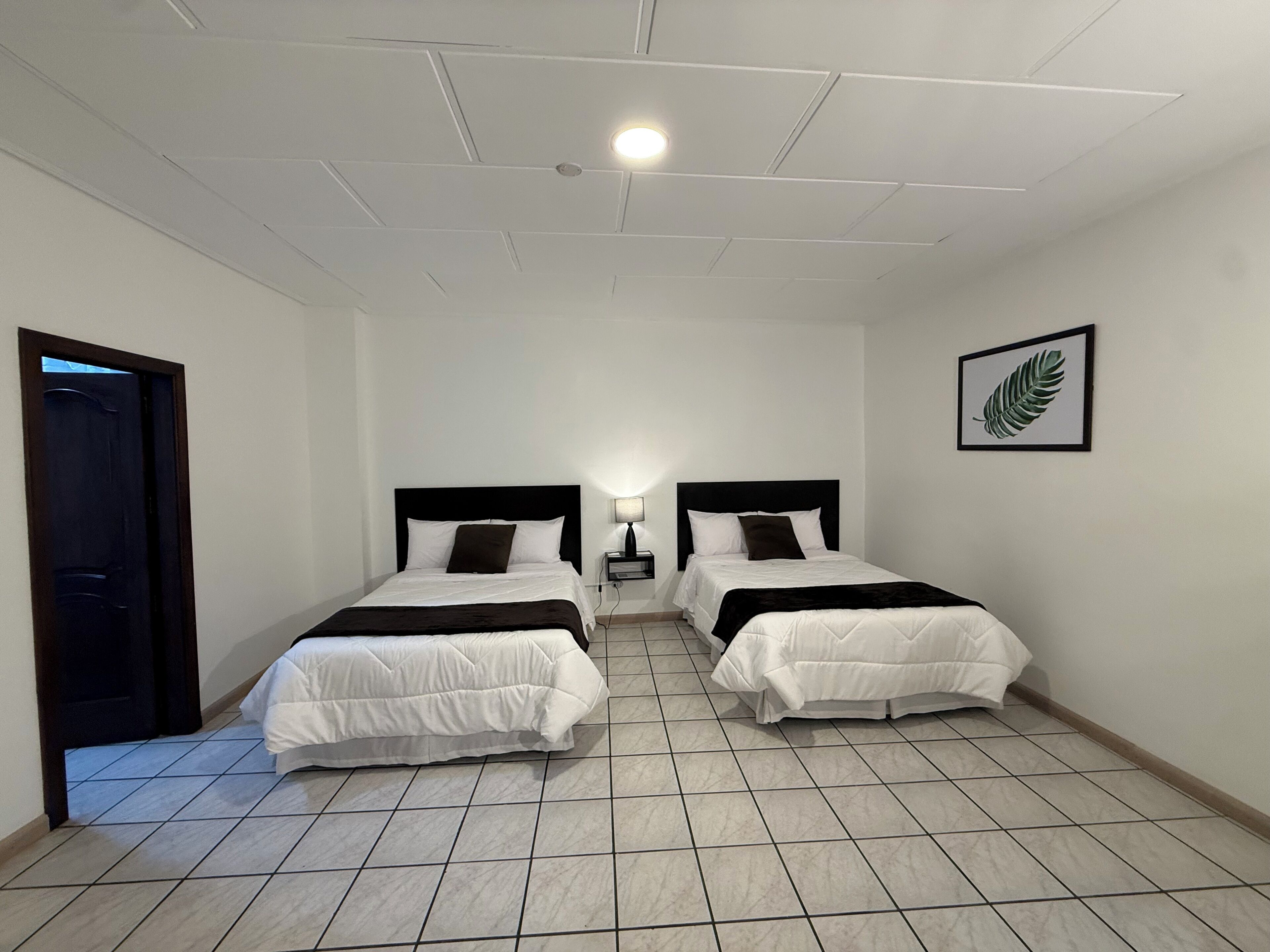 Habitación de hotel con dos camas, una puerta, una mesita de noche, una lámpara y una hoja enmarcada en la pared.