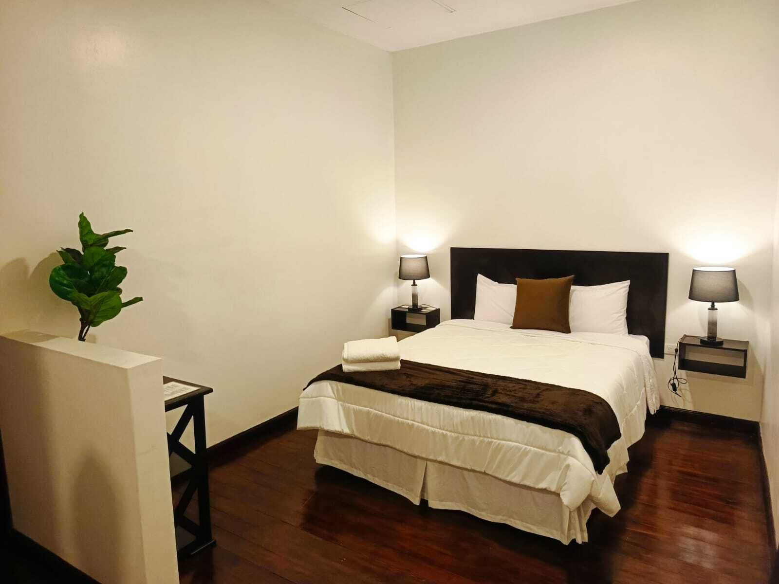 Habitación de hotel con cama, dos mesitas de noche con lámparas, una planta y una pared blanca.