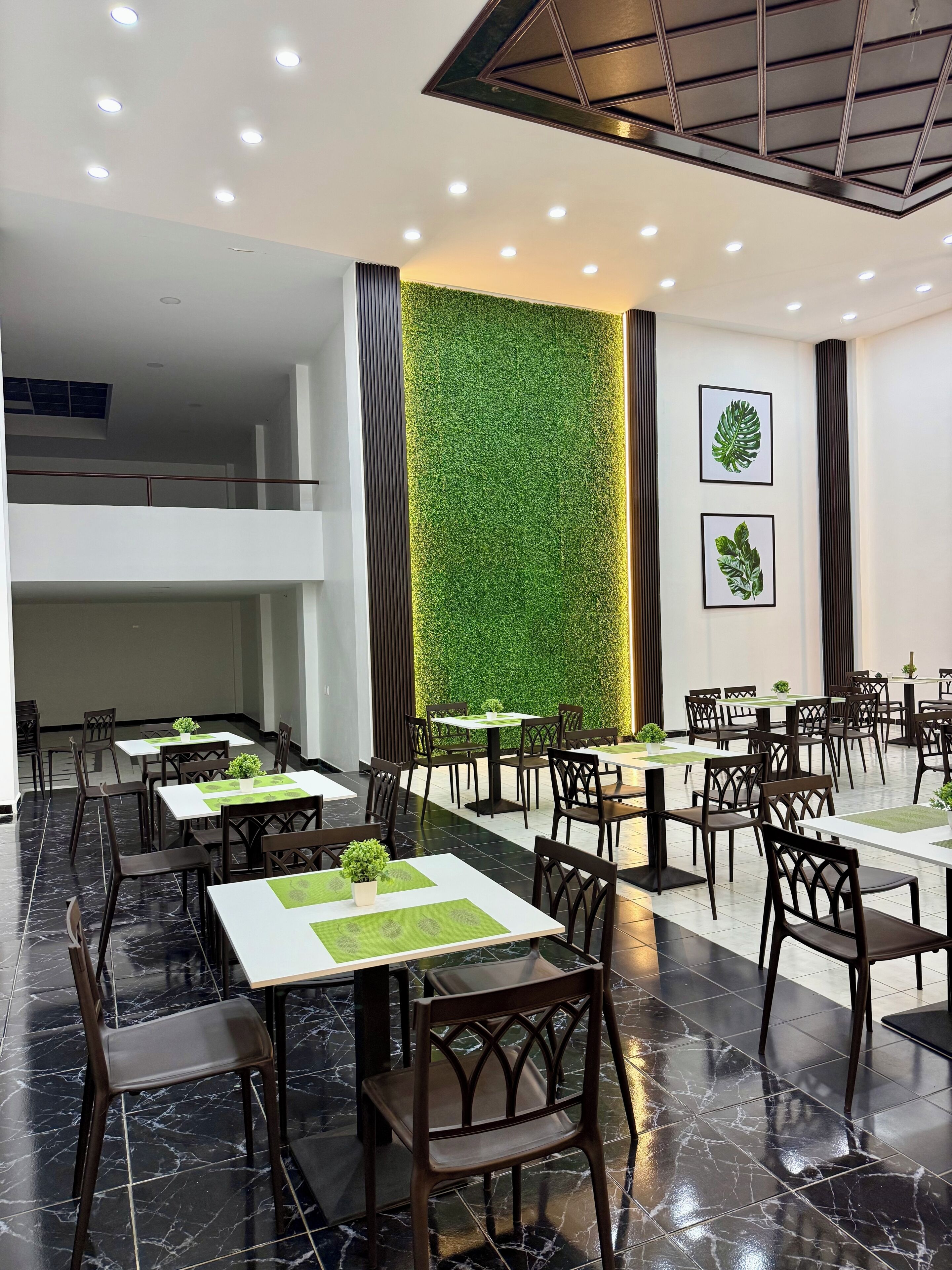 Un área de comedor moderna con mesas y sillas, una pared verde con plantas e iluminación empotrada en el techo.