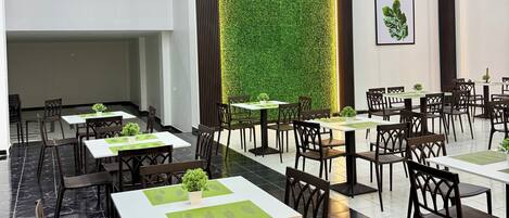 Un área de comedor moderna con mesas y sillas, una pared verde con plantas e iluminación empotrada en el techo.
