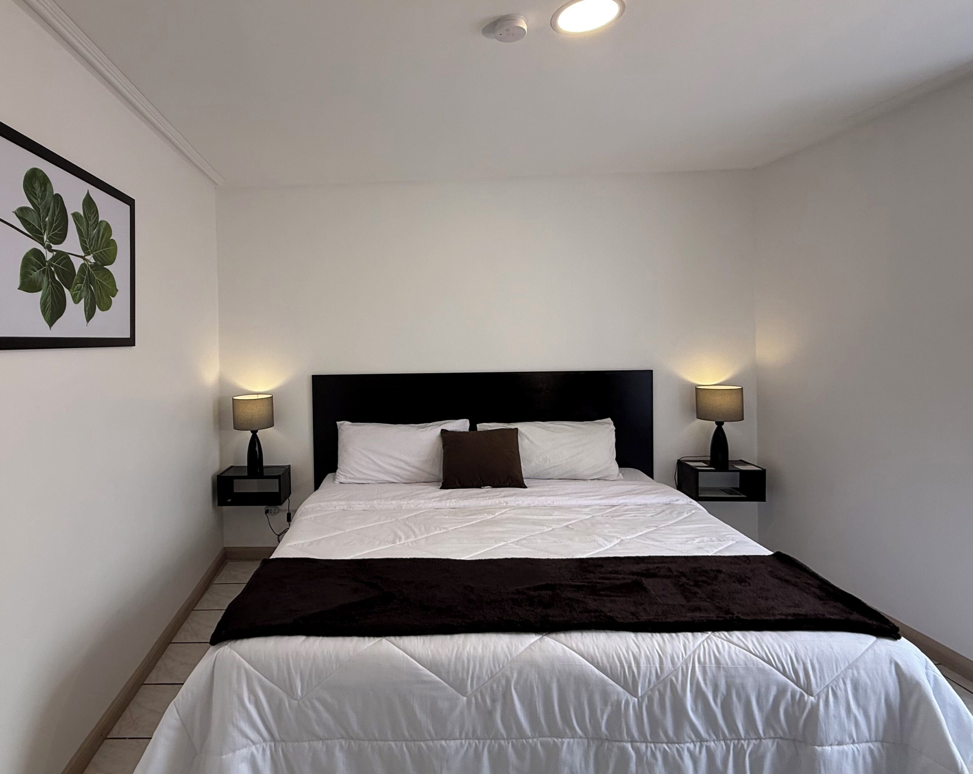 Una cama cuidadosamente tendida con ropa blanca y un cabecero oscuro, flanqueada por dos mesitas de noche con lámparas, en una habitación con paredes blancas y una luz de techo.