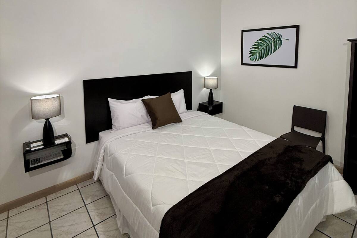 Une chambre à coucher comprenant un lit, deux tables de chevet avec des lampes, un tableau accroché au mur et une chaise.