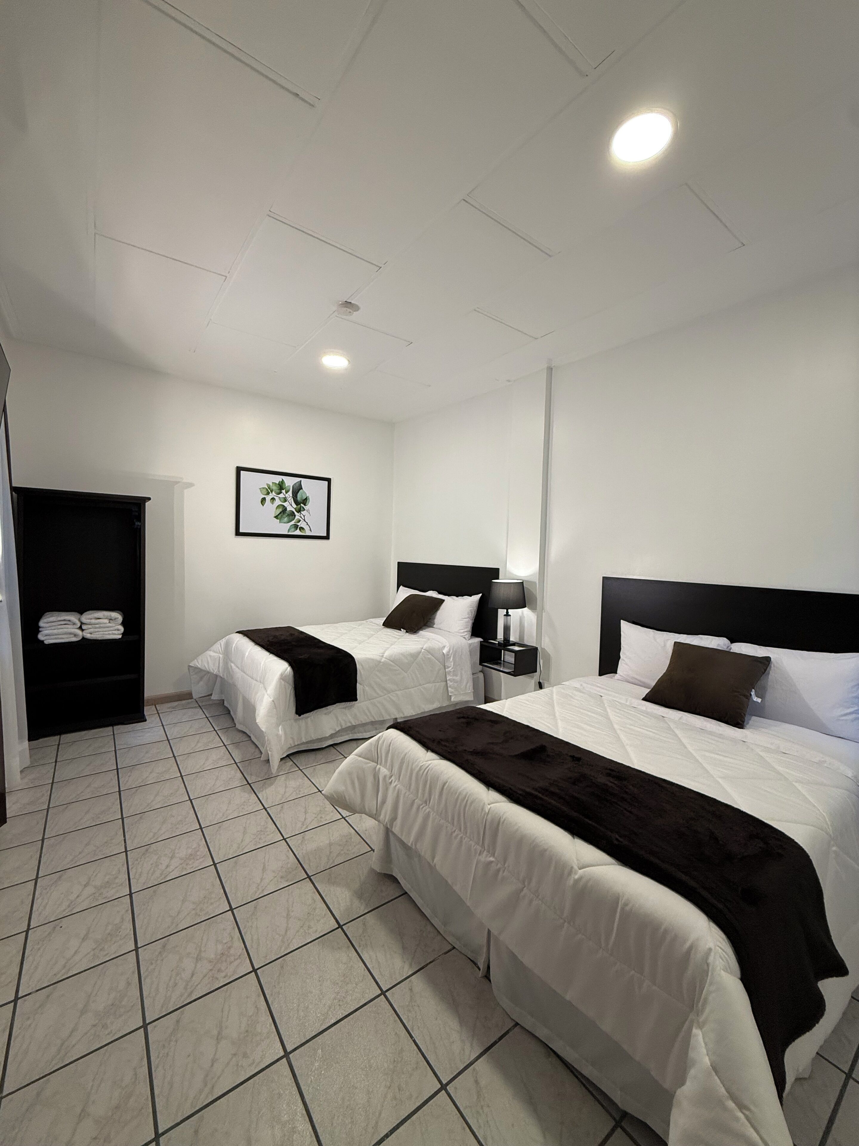 A hotel room with two beds, a wardrobe, a nightstand, a lamp, and a framed picture on the wall.