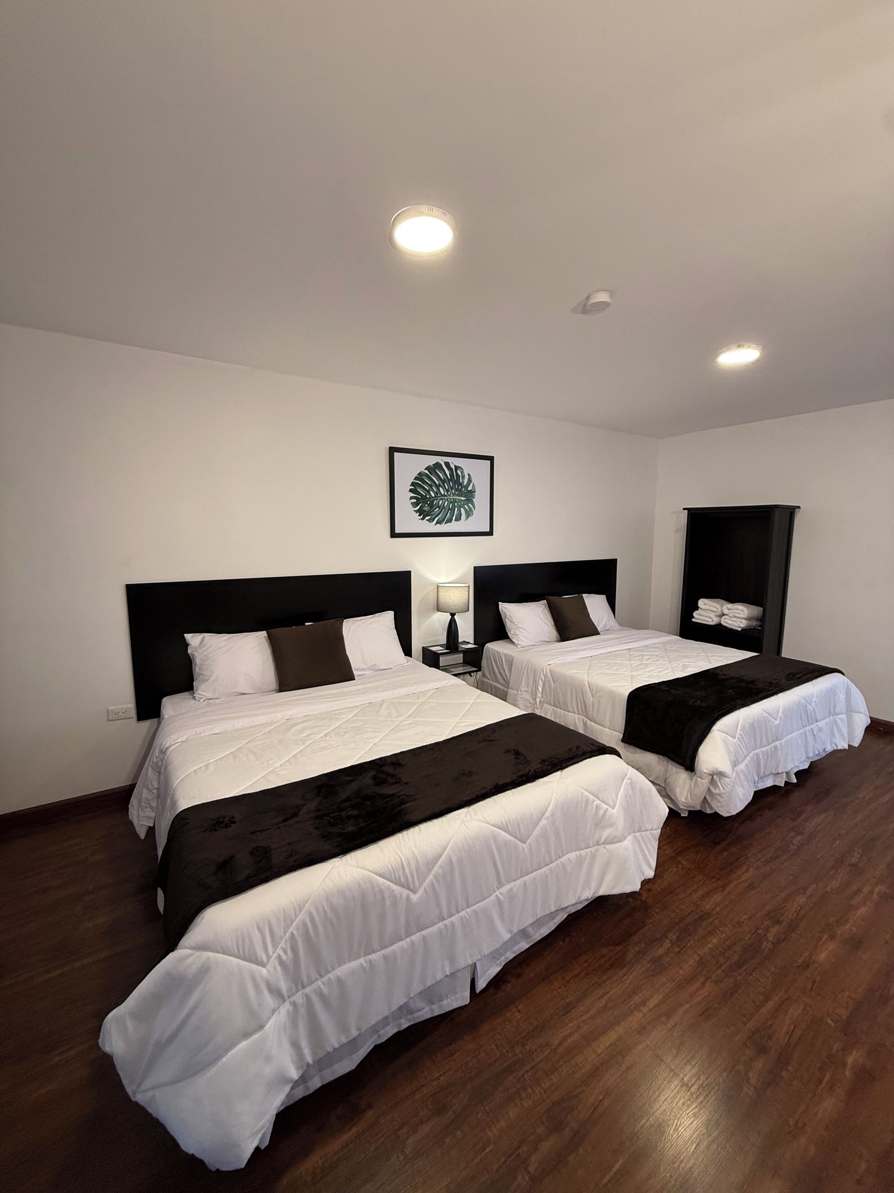 Habitación de hotel con dos camas, una mesita de noche, una lámpara, una obra de arte con hojas enmarcadas y un armario.