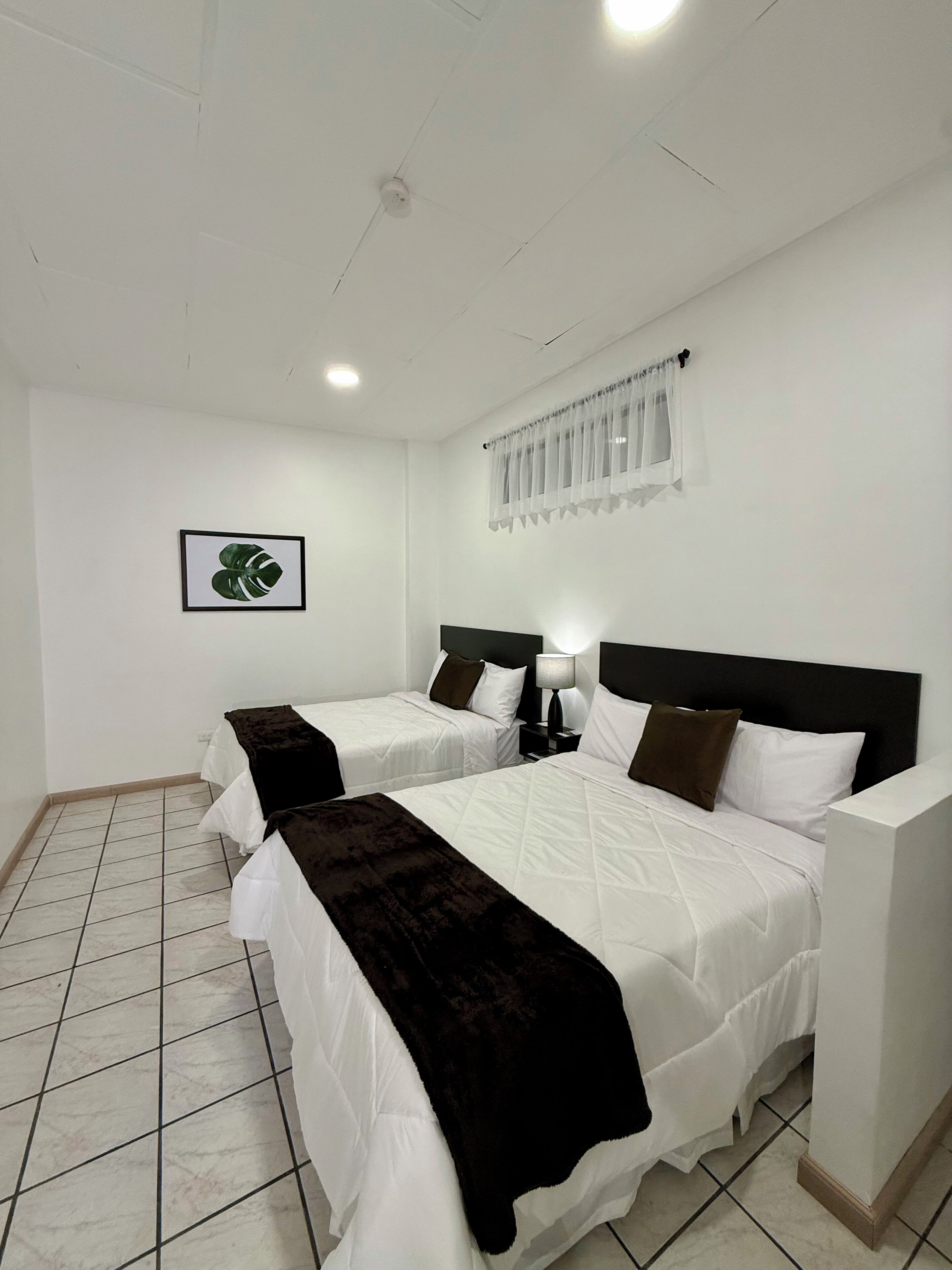 A hotel room with two beds, a ceiling fan, a window with sheer curtains, and a framed picture on the wall.