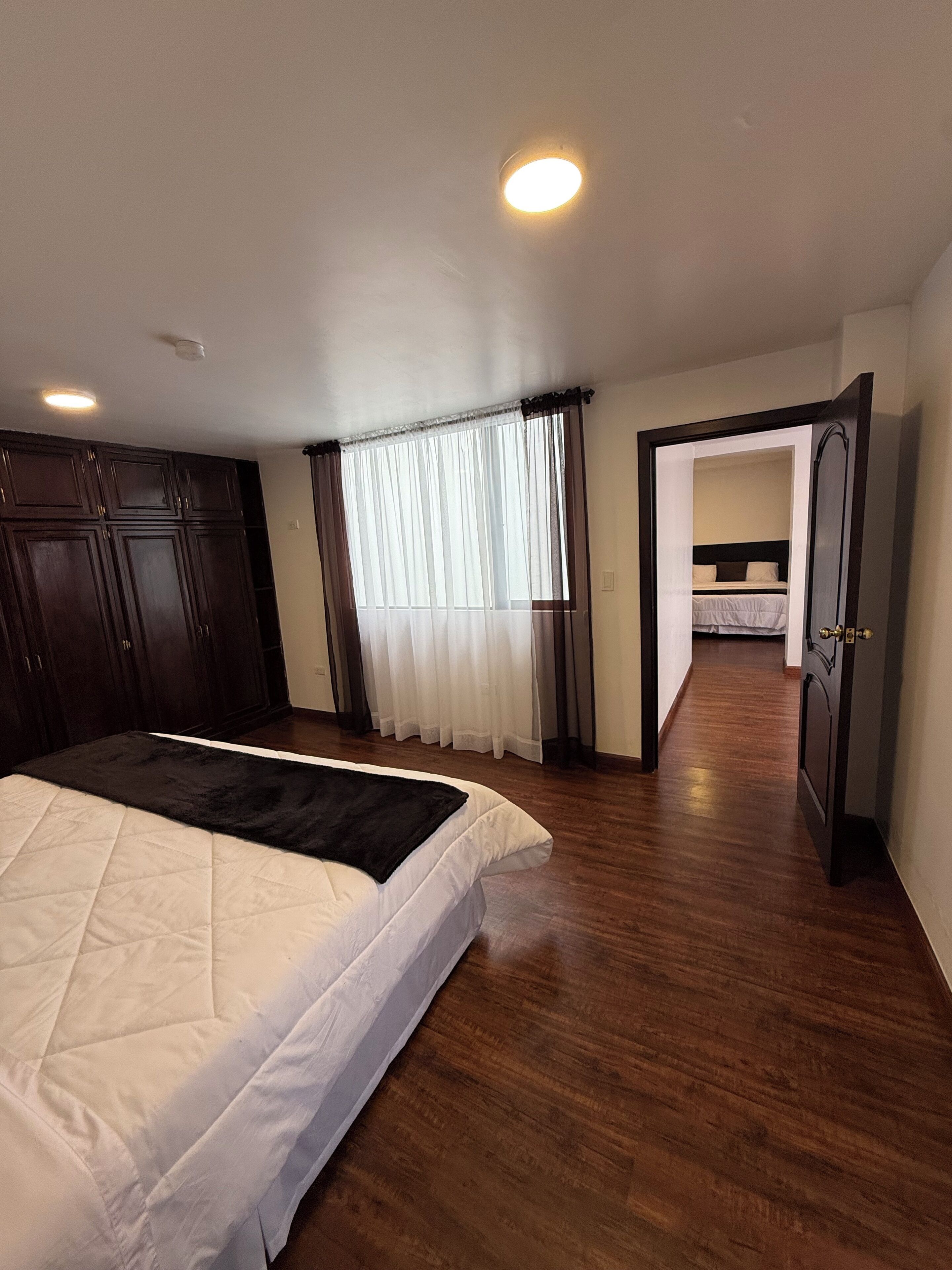 A bedroom with a bed, a wardrobe, a window with curtains, and a door leading to another room.