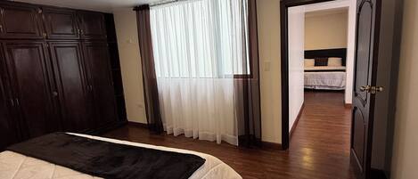 A bedroom with a bed, a wardrobe, a window with curtains, and a door leading to another room.
