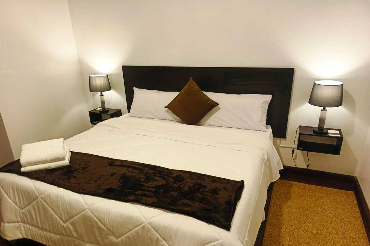 Un lit bien fait, avec une tête de lit, deux tables de chevet avec des lampes, une serviette blanche pliée et un plaid marron.