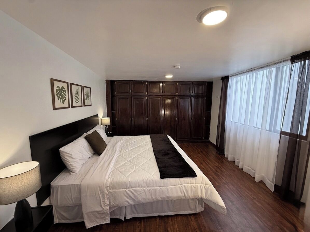 A bedroom with a bed, a nightstand, a wall-mounted lamp, a dark headboard, a large window with curtains, and a ceiling light.
