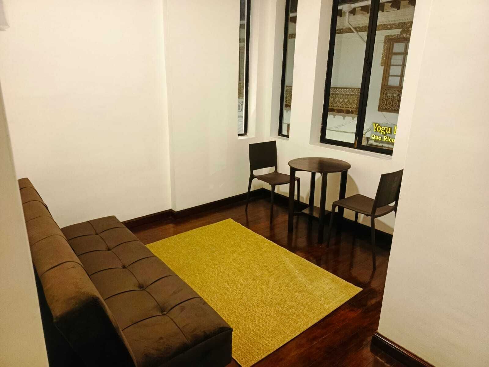 Una habitación con un sofá marrón, un tapete amarillo, una mesita redonda y dos sillas.