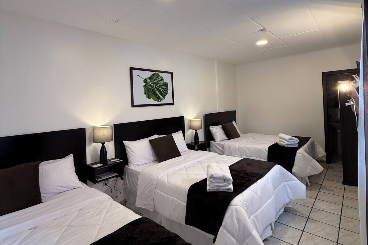 Une chambre d’hôtel avec trois lits, un ventilateur de plafond, une plante décorative dans un cadre et un sol carrelé.