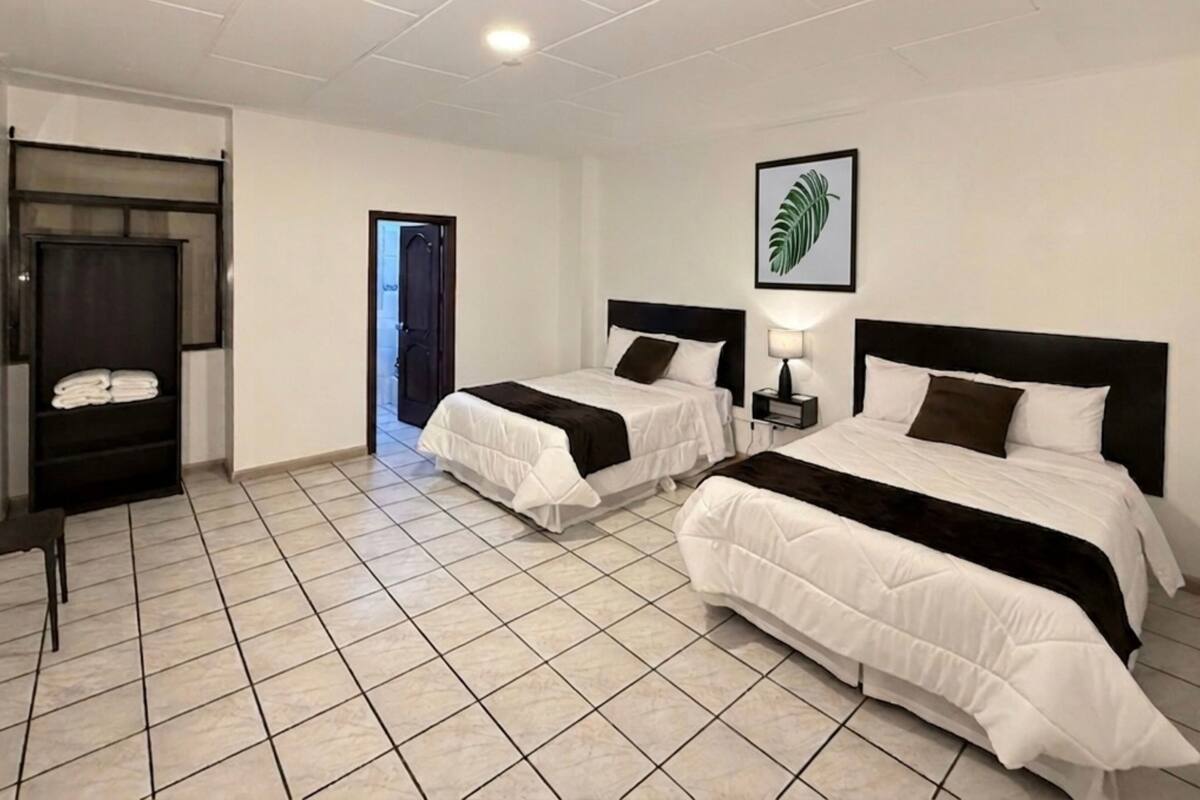 Une chambre d’hôtel avec deux lits, une armoire, une porte donnant sur une autre pièce et une œuvre d’art représentant une feuille, encadrée, accrochée au mur.