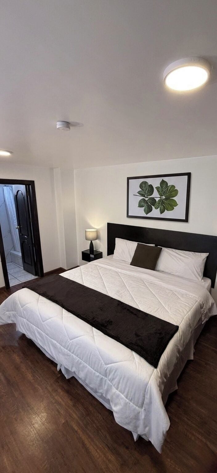 Een slaapkamer met een bed, nachtkastje, een wandlamp en een lijst met een plant.